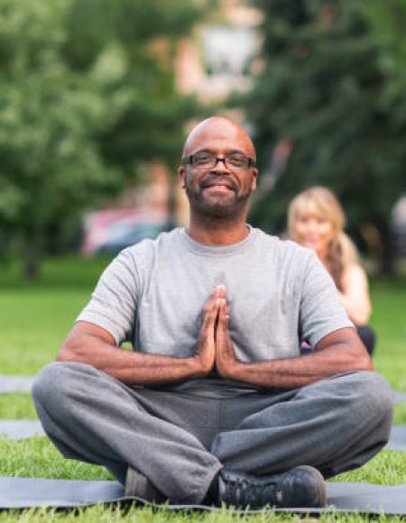 A multi-ethnic group of seniors is attending a yoga class outdoors. The group is sitting on yoga mats. They are meditating. The individual in focus is a black man. He is sitting at the front of the group. He is smiling directly at the camera.